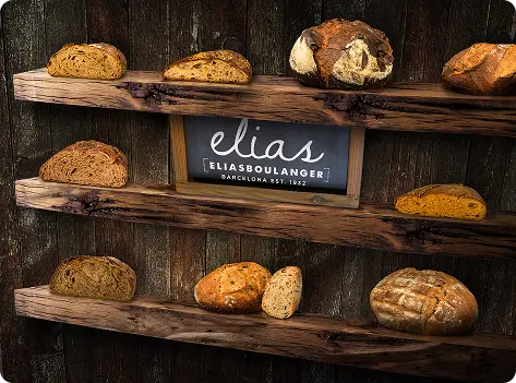 Verschiedene Brotsorten auf Holzregalen in einer Bäckerei, mit dem Schriftzug "Elias Boulanger" und dem Gründungsjahr 1962 in Barcelona.