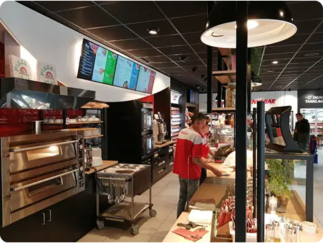 Moderne Restaurantküche mit Pizzaofen und Menütafeln, während ein Mitarbeiter Lebensmittel zubereitet.