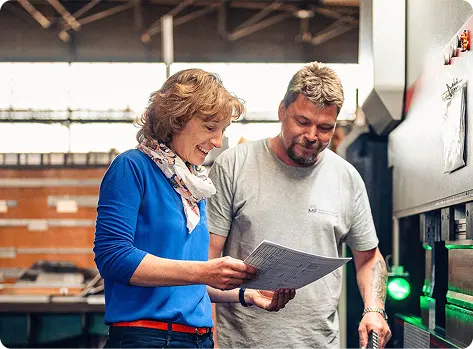 Zwei Personen in einer Fabrik besprechen technische Unterlagen. Eine Frau mit blonden Locken und blauem Pullover lächelt, während sie mit einem Mann in grauem T-Shirt spricht. Im Hintergrund sind Maschinen und industrielle Elemente sichtbar.