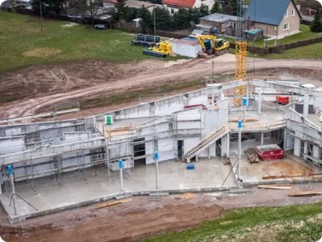 Baufortschritt eines modernen Wohnhauses mit Betonfundament, Gerüsten und einem Kran im Hintergrund, umgeben von unbebautem Gelände und angrenzenden Grundstücken.