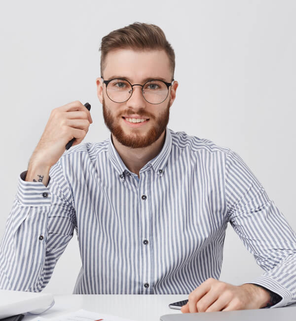 Junger Mann mit Brille und gestreifter Hemd sitzt am Schreibtisch, hält einen Stift in der Hand und lächelt. Auf dem Tisch sind Notizen und Bürobedarf sichtbar.