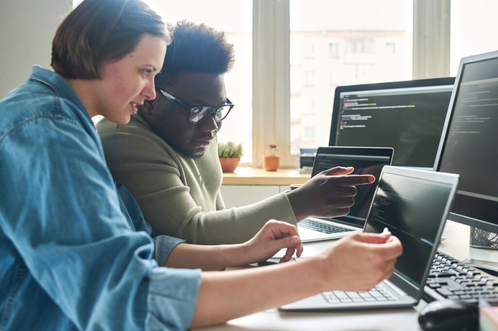Zwei Personen arbeiten gemeinsam an Computerprogrammen, während sie auf mehrere Bildschirme mit Code schauen. Der Raum ist hell erleuchtet und es sind verschiedene Laptops und Computer sichtbar.